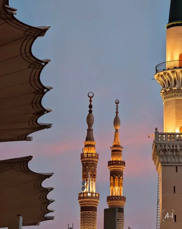 Two lit minarets between a canopy and a tower against a pink evening sky