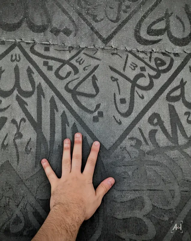 A hand resting on the black kiswa, its woven calligraphy close up
